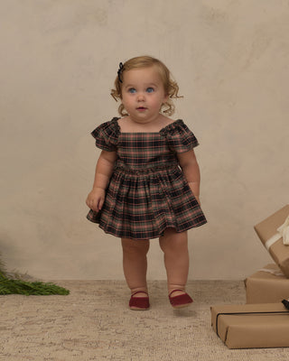 Noralee Margot Romper - Tartan
A toddler girl wearing a dark brown and beige plaid dress with puff sleeves, standing against a textured beige backdrop with wrapped gift boxes on the floor.