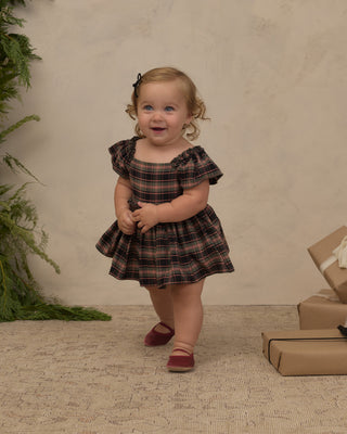 Noralee Margot Romper - Tartan
A toddler girl wearing a dark brown and black plaid off-the-shoulder dress and burgundy shoes, standing in a studio setting with a textured beige backdrop, greenery on the left, and wrapped gifts on the right.