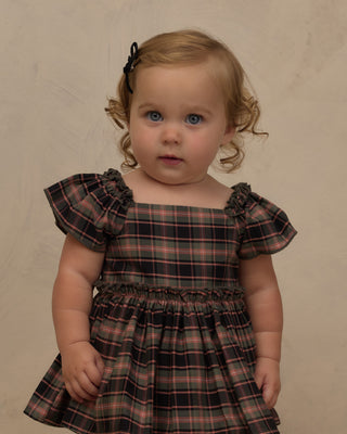 Noralee Margot Romper - Tartan
A toddler girl with blonde curly hair and blue eyes wearing a dark plaid dress with ruffled flutter sleeves, standing against a neutral beige studio backdrop.