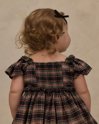Noralee Margot Romper - Tartan
A toddler viewed from behind wearing a smocked plaid dress in dark green, red, and black, with a black bow in her hair, set against a neutral textured beige background.