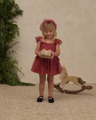 Noralee Gigi Tutu - Ruby 
A young girl with blonde hair wearing a dusty rose pink tulle dress and matching headband, holding a small gift box. She is standing in front of a textured beige wall with a wooden rocking horse and green foliage in the background.