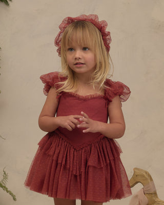 Noralee Gigi Tutu - Ruby 
A young blonde girl wearing a dusty rose red tiered tulle dress with ruffled cap sleeves and a matching ruffled headband, standing against a textured beige wall.
