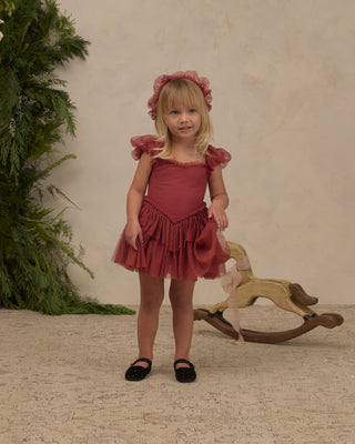 Noralee Gigi Tutu - Ruby 
A young girl with blonde hair stands wearing a dusty rose pink ruffled dress with puff sleeves and a matching headband, standing in front of a textured beige wall with a wooden rocking horse and green foliage in the background.