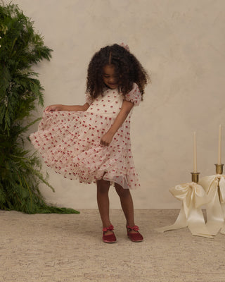 Noralee Dottie Dress - Ruby Bows
A young girl with curly dark hair wearing a light pink short-sleeve dress with a red heart pattern and red shoes, twirling to show off the dress. She is standing in a styled studio setting with a textured beige wall, a green plant on the left, and a cream ribbon and candle holder prop on the right.