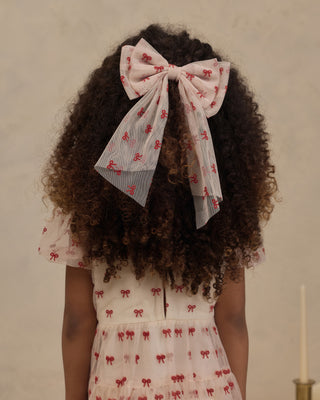Noralee Alice Bow - Ruby Bows
A child viewed from behind wearing a large sheer bow headband with a red bow print and a matching short-sleeve dress with small red bow motifs, set against a plain beige wall.