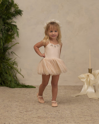 Noralee Clementine Tutu - Pearl
A young girl with blonde hair wearing a pale peach-beige tutu dress with lace detailing on the bodice, a matching headband, and ballet slippers, posing in a studio setting with a textured beige wall and green foliage.