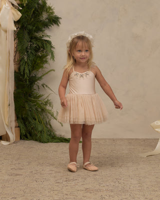 Noralee Clementine Tutu - Pearl
A young girl with blonde hair wearing a pale peach-beige tutu dress with a matching floral headband and pink ballet slippers, standing in a studio setting with a textured beige wall and green foliage backdrop.