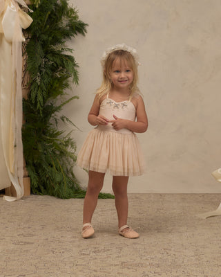 Noralee Clementine Tutu - Pearl'
A young girl with blonde hair stands in a studio wearing a pale blush pink sleeveless tutu dress with a floral lace bodice, a matching flower crown, and ballet flats. She is posed in front of a textured beige wall with a green foliage prop on the left.