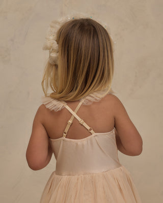 Noralee Clementine Tutu - Pearl
A young girl viewed from behind wearing a pale blush pink dress with ruffled straps and a matching ruffled hair accessory against a soft beige textured background.