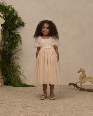 Noralee Dorothy Dress - Pearl
A young girl with curly dark hair stands wearing a two-piece formal dress with a cream lace bodice and puff sleeves, paired with a light peach tulle skirt. She is wearing gold shoes and a matching hair bow, standing in a styled room with a textured beige wall, green foliage, and a wooden rocking horse.