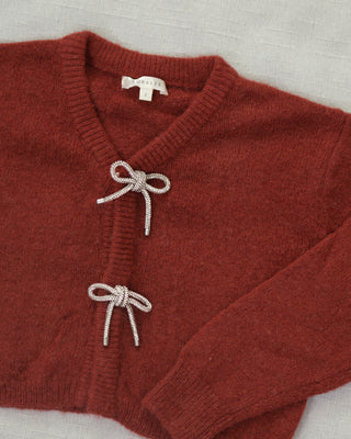 Noralee Bow Cardigan - Ruby
A close-up of a rust-red knit cardigan sweater with two decorative knotted bow details on the front, laid flat on a light textured fabric background. The brand name 'SARAH' is visible on the inner neck label.