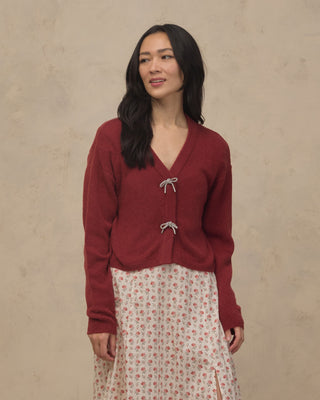 Noralee Women's Bow Cardigan - Ruby
Woman wearing a red cardigan over a floral skirt against a beige background