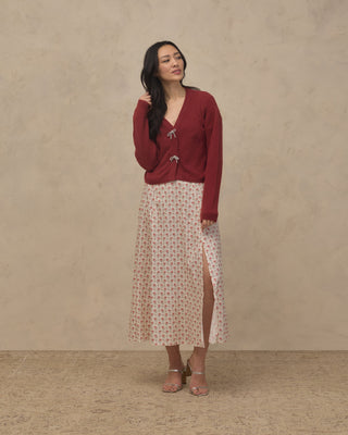 Noralee Women's Bow Cardigan - Ruby
A woman stands against a textured beige studio wall wearing a dark red long-sleeve cardigan with decorative buttons over a cream midi skirt with a small repeating floral pattern and a front slit, paired with tan heeled sandals.