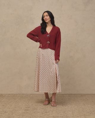 Noralee Women's Bow Cardigan - Ruby
A woman stands against a textured beige studio backdrop wearing a burgundy cardigan with drawstrings and a light pink patterned midi skirt with a side slit, paired with clear high-heeled sandals.