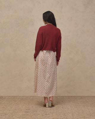 Noralee Women's Bow Cardigan - Ruby
A woman is shown from behind wearing a dark red long-sleeve knit top and a cream-colored midi skirt with a small red floral pattern, standing against a textured beige wall.