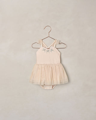 Noralee Clementine Tutu - Pearl
A baby girl's sleeveless bodysuit in a soft cream or light beige color, featuring delicate floral embroidery on the chest and a tiered tulle tutu skirt. The bodysuit is displayed on a metal hanger against a textured, warm beige wall.