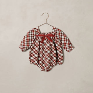 Noralee Annette Romper -  Holiday Tartan
A baby romper in a red and white gingham pattern, featuring a red bow at the neckline, hanging on a metal hanger against a textured beige wall.