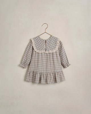 Noralee Noelle Dress - Fall Plaid
A toddler girl's long-sleeve dress in a beige and gray plaid pattern, featuring a large white lace Peter Pan collar and a ruffled hem, hanging on a wooden hanger against a textured beige wall.