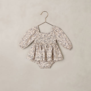 Noralee Elodie Romper - Blue Garden
A baby long-sleeve romper with a smocked bodice and ruffled hem, featuring a delicate beige and gray floral pattern, hanging on a metal hanger against a textured beige wall.