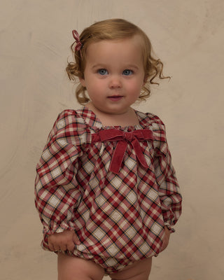 Noralee Annette Romper -  Holiday TartanA toddler girl with curly light brown hair wearing a red and cream plaid long-sleeve romper with a red bow at the neckline, standing against a neutral beige textured backdrop.