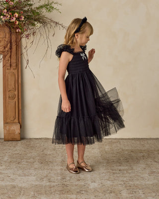 Noralee Clara Dress - Black
Young girl in a black dress standing against a beige wall with decorative elements.