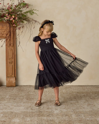 Noralee Clara Dress - Black 
Young girl in a black dress with a bow, standing in a room with a fireplace and floral decorations.