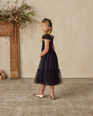 Noralee Clara Dress - Black
Young girl in a black dress standing in a room with a fireplace and floral decorations.