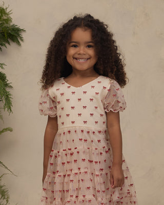 Noralee Dottie Dress - Ruby Bows
A young girl with curly brown hair smiling, wearing a short-sleeved cream-colored dress with a red bow pattern, standing against a textured beige wall.