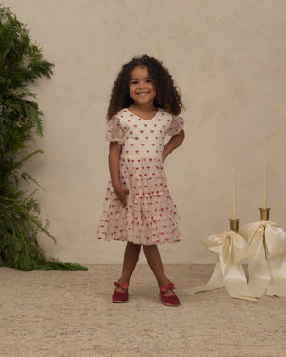 Noralee Dottie Dress - Ruby Bows 
A young girl with curly dark hair stands in a studio setting wearing a short-sleeve pink dress with a red pattern, paired with red shoes. The background features a textured beige wall, a green plant on the left, and decorative cream ribbons and candle holders on the right.
