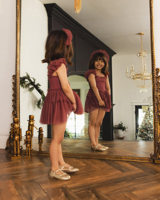 Noralee Gigi Tutu - Ruby
A young girl stands in front of a large ornate mirror wearing a dusty rose pink ruffled tutu dress and matching headband, with her reflection visible. The setting is a classic room with a herringbone wood floor, gold mirror frame, and a chandelier.