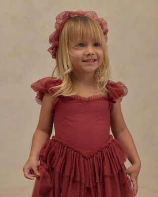 Noralee Gigi Tutu - Ruby
A young blonde girl wearing a dusty rose red tiered dress with ruffled sleeves and a matching ruffled headband, standing against a neutral beige background.