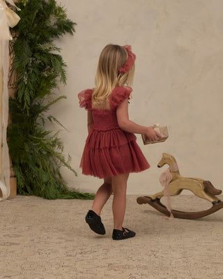 Noralee Gigi Tutu - Ruby
A young girl with blonde hair is seen from behind wearing a dusty rose pink tiered tulle dress with ruffled sleeves and a matching headband. She is walking on a textured beige floor next to a wooden rocking horse and green foliage against a neutral beige wall.