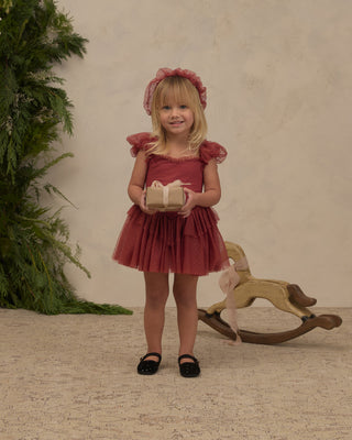 Noralee Gigi Tutu - Ruby
A young girl stands in a studio wearing a dusty rose pink tulle dress with ruffled sleeves and a matching headband, holding a small wrapped gift box. A wooden rocking horse and green foliage are visible in the background against a textured beige wall.