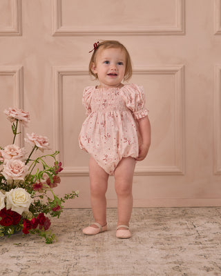 Noralee Hazel Romper - Bouquet
Baby in a floral romper standing next to flowers against a beige paneled wall.