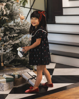 Noralee Janette Dress - Winter Ditsy
A young girl wearing a black short-sleeve dress with a red bow headband and red shoes, standing in a festive indoor setting next to a decorated Christmas tree and wrapped gifts.
