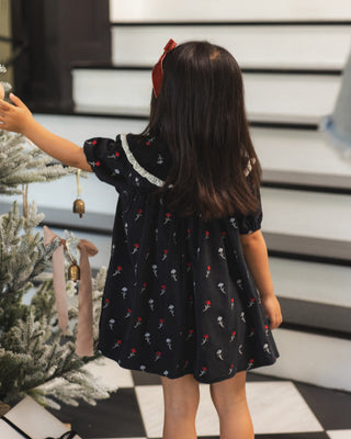 Noralee Janette Dress - Winter Ditsy
A young girl viewed from behind wearing a black short-sleeve dress with a red and white skull pattern, standing next to a decorated Christmas tree in front of white steps.
