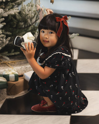 Noralee Janette Dress - Winter Ditsy
A young girl with a red bow in her hair crouches in front of a Christmas tree, holding a smartphone to take a photo. She is wearing a black dress with a white ruffle trim and small red and white skeleton print, paired with red shoes.