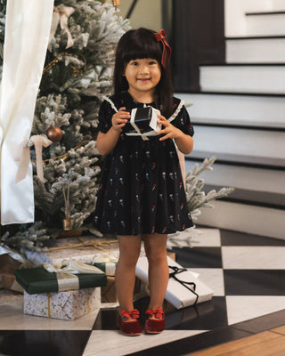 Noralee Janette Dress - Winter Ditsy 
A young girl stands in front of a decorated Christmas tree wearing a black short-sleeve dress with a small red and white pattern, holding a gift box. She is wearing red shoes, with wrapped presents on the floor and a black-and-white checkered floor beneath her.
