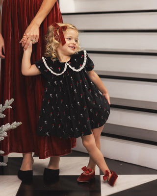 Noralee Janette Dress - Winter Ditsy
A young girl wearing a black short-sleeve dress with a white ruffled Peter Pan collar and a red cherry print, walking while holding an adult's hand. The adult is wearing a dark red velvet dress.