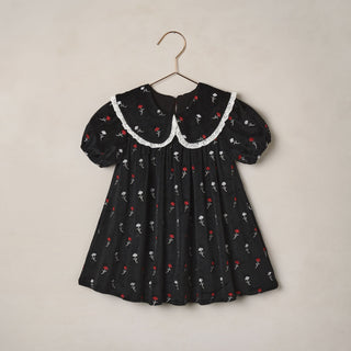 Noralee Janette Dress - Winter Ditsy
A black short-sleeve baby dress with a white lace Peter Pan collar, featuring a delicate red and white floral print, hanging on a gold-colored metal hanger against a warm beige background.