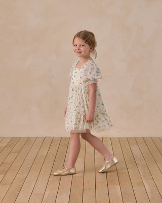 Noralee Louella Dress - Bows
Young girl in a floral dress standing on a wooden floor with a beige wall background