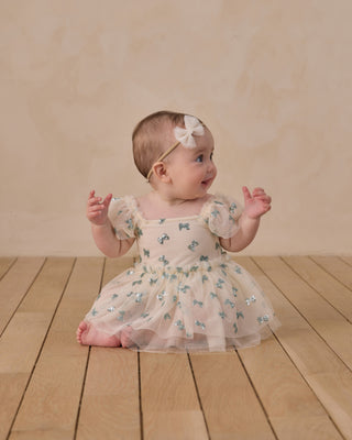 Noralee Margot Romper - Bows\
Baby in a floral dress standing on a wooden floor with a beige background