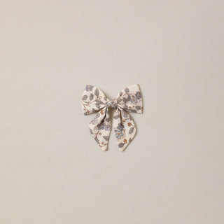 Noralee Norah Bow - Blue Garden
A small fabric bow accessory with a delicate floral print in muted blue, brown, and gray tones on a light cream background, centered against a solid light beige backdrop.
