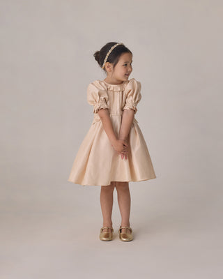 Noralee Pearl Headband - Ivory
Young girl wearing a beige dress with puffed sleeves against a plain background