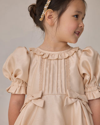 Noralee Pearl Headband - Ivory
Young girl wearing a beige dress with ruffled details and a bow on a plain background