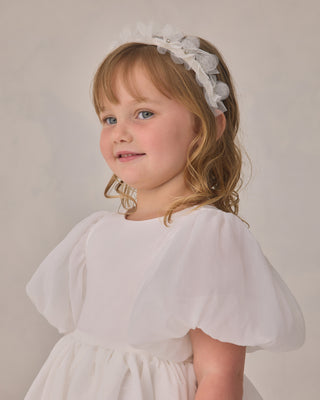 Noralee Pixie Headband - Ivory
Young girl wearing a white dress with a bow headband against a plain background