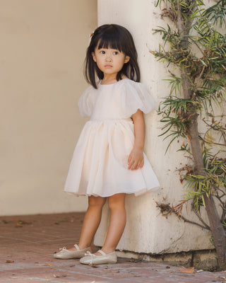Noralee Sofia Dress - Blush
Young girl in a white dress standing next to a columned building with greenery.