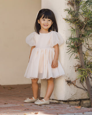 Noralee Sofia Dress - Blush
Young girl in a white dress standing against a beige wall with greenery.