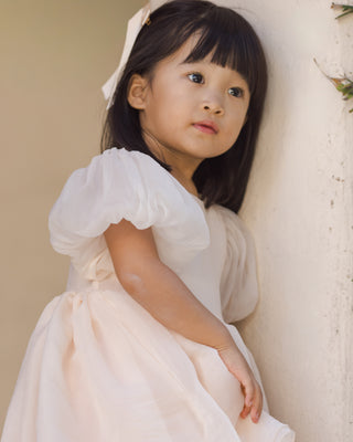 Noralee Sofia Dress - Blush
Young girl in a white dress with puffed sleeves against a neutral background