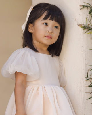 Noralee Sofia Dress - Blush
Young girl in a white dress with puffed sleeves standing against a neutral background
